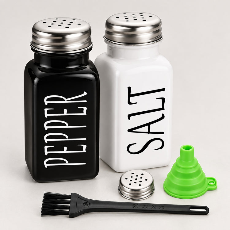 A set of two ceramic shakers, one black labeled 'PEPPER' and one white labeled 'SALT', with a small green funnel, a metal shatter guard, and a black cleaning brush on a light gray background.