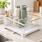 A white plastic and wire dish drying rack with light wood handles, shown on a kitchen counter with mugs, bottles, glasses, and utensils inside.