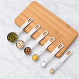 Six steel measuring spoons holding grains and spices, arranged beside a wooden board.