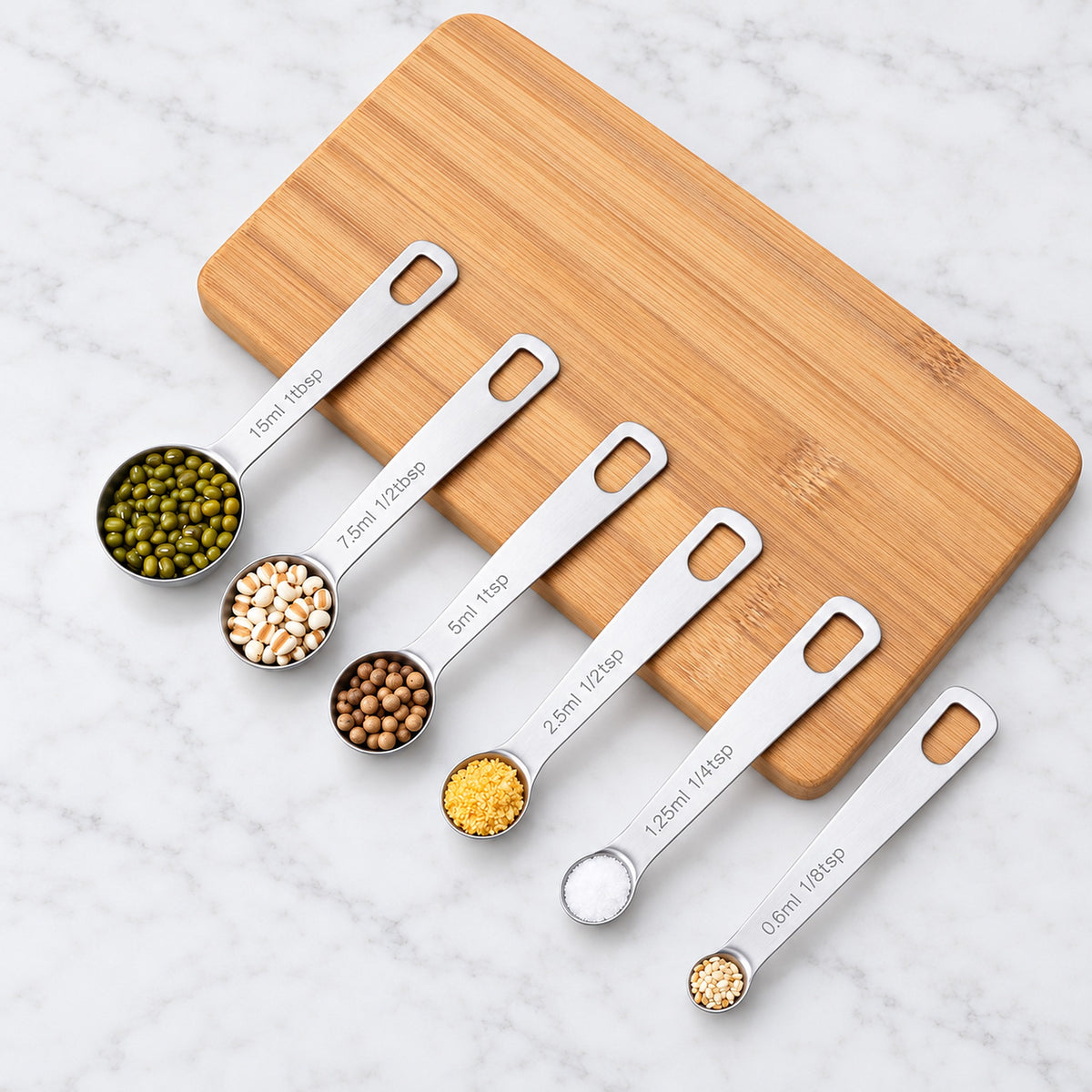 Six steel measuring spoons holding grains and spices, arranged beside a wooden board.
