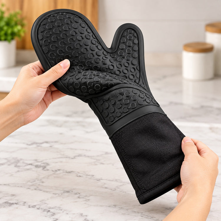 A black silicone oven mitt with textured non-stick and grease-repellent surfaces, being held by two hands over a light gray marble kitchen countertop.