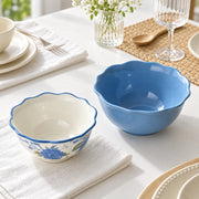 Two ceramic bowls on a styled dining table: one blue bowl with a scalloped rim and one white bowl with a blue floral pattern and matching blue scalloped rim. The scene includes plates, napkins, a glass vase with flowers, a wine glass, and a wooden spoon.