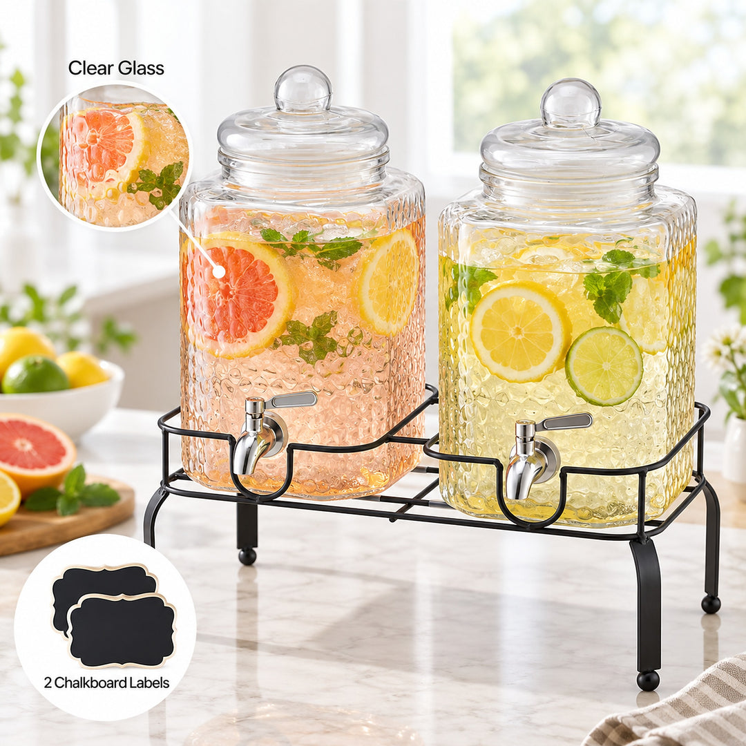 Two clear glass beverage dispensers with lids and metal spouts, shown on a black stand on a kitchen counter. The left dispenser contains a pink drink with grapefruit and mint, and the right dispenser contains a yellow drink with lemon and lime.