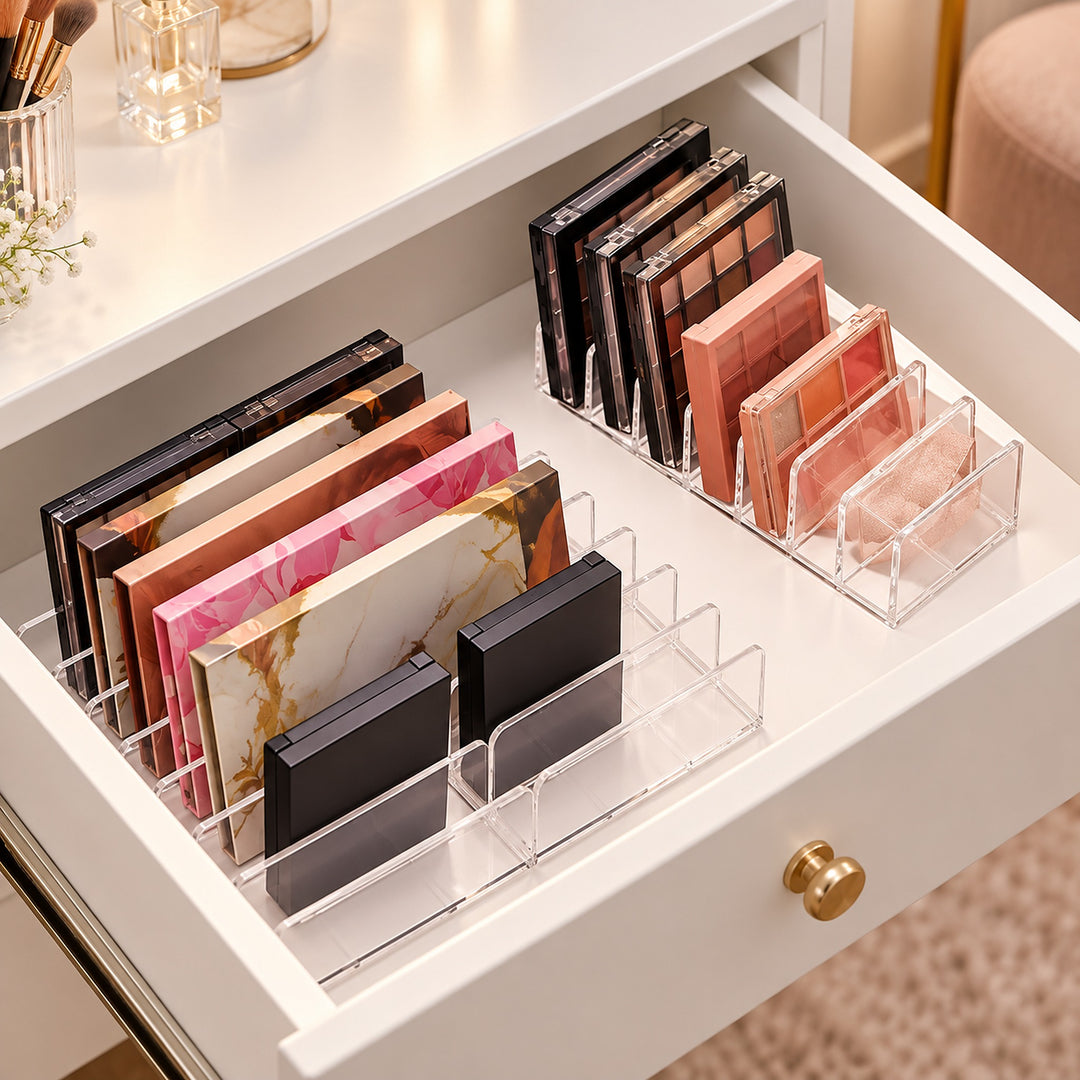 Clear acrylic makeup organizer trays arranged inside an open white drawer, holding multiple eyeshadow palettes and compact powders in neutral, pink, and gold tones.