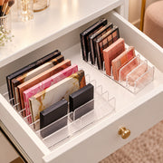 Clear acrylic makeup organizer trays arranged inside an open white drawer, holding multiple eyeshadow palettes and compact powders in neutral, pink, and gold tones.