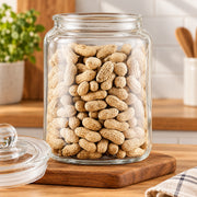 A clear glass jar filled with peanuts in their shells, placed on a wooden cutting board on a kitchen counter with the glass lid resting beside it.