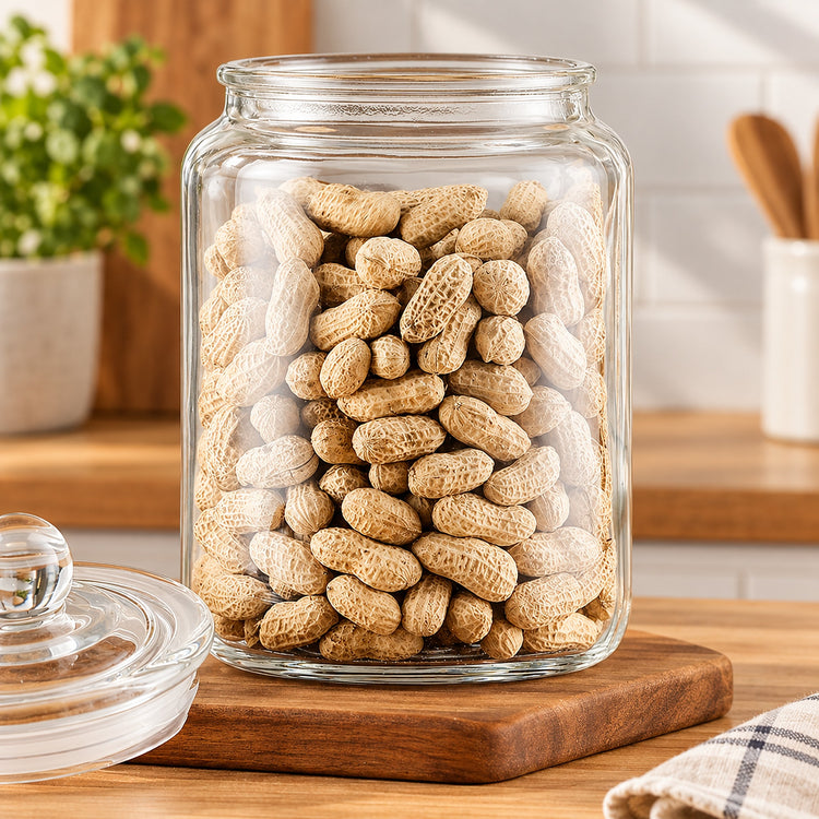 A clear glass jar filled with peanuts in their shells, placed on a wooden cutting board on a kitchen counter with the glass lid resting beside it.