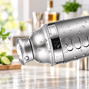 A close-up of a stainless steel cocktail shaker covered in water droplets, set against a softly blurred kitchen background with mint, lime, and glassware.