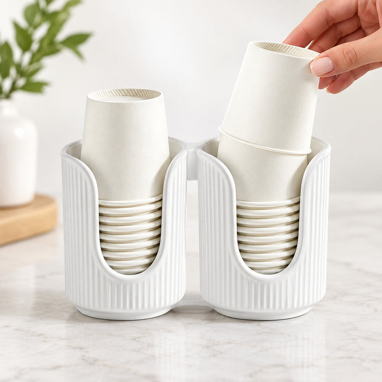 A white ribbed dual-cup holder on a light marble surface, with two stacks of off-white paper cups inside and a hand placing another cup into the right slot.