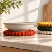 A white ceramic bowl resting on a round orange felt coaster on a light marble countertop, with a stack of colorful felt coasters in yellow, green, and white to the right.