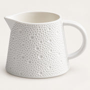 A white ceramic pitcher with a textured, dimpled surface pattern and a curved handle, shown on a light cream background.