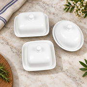 Three white ceramic butter dishes with matching lids arranged on a light marble surface, styled with a folded cloth, flowers, and green leaves.
