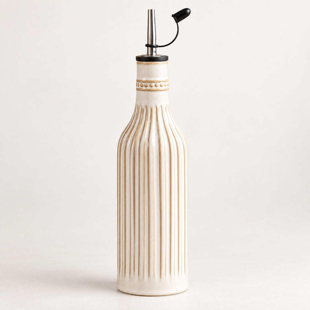 A ribbed ceramic oil bottle in cream with gold detailing, featuring a black pour spout with a metal rod and lever, set against a plain light gray background.
