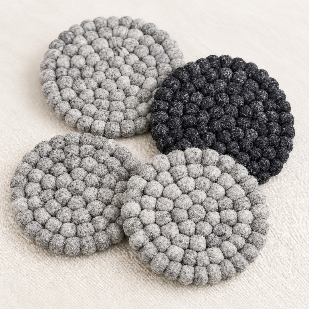 Four round felt coasters with a textured bumpy knit pattern, shown in three shades of gray and one black, arranged on a light beige fabric background.