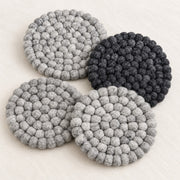 Four round felt coasters with a textured bumpy knit pattern, shown in three shades of gray and one black, arranged on a light beige fabric background.