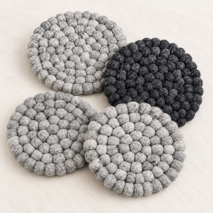 Four round felt coasters with a textured bumpy knit pattern, shown in three shades of gray and one black, arranged on a light beige fabric background.