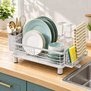 A white plastic and wire under-sink dish rack on a light wood kitchen countertop, shown holding plates, bowls, wine glasses, utensils, and a striped towel. A sink is visible on the right, with a window and plants in the background.