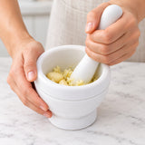 White mortar and pestle crushing peeled garlic in a kitchen prep scene.