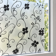 Black floral and scroll vine window film applied to a frosted glass window pane, with a white window frame and blurred green outdoor foliage visible in the background.