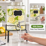 Clear glass lemonade dispenser with a black metal stand, filled with lemons, ice, and mint, with a faucet pouring into a glass. A hand is visible holding the glass, and an inset shows the stand and text 'With Stand Easy Cup Clearance'.