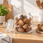 A clear glass storage jar filled with walnuts sits on a wooden countertop, with the matching glass lid leaning beside it. The scene is styled in a bright kitchen with a cutting board, plant, utensils, and a small bowl of walnuts visible in the background.