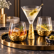 Three gold hammered glass drinking vessels on a round tray: a martini glass with olives, an rocks glass with ice and garnish, and a tumbler with a lemon slice and herb, set on a dark marble surface.