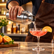 A bartender in a brown apron pours a pink cocktail into a coupe glass with an orange slice garnish, with oranges and mint on a counter in the foreground and a blurred bar background.
