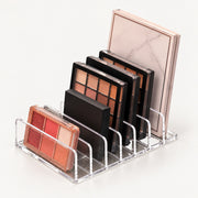 A clear acrylic makeup organizer stand holding several eyeshadow palettes in different colors and sizes against a clean white background.