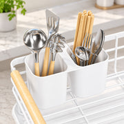A white dual-compartment utensil holder filled with metal kitchen tools and chopsticks, placed on a white wire rack in a bright kitchen setting.