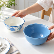 Two ceramic bowls with scalloped edges are shown on a white table. One bowl is held by hands and is decorated with blue floral patterns on a white interior and exterior, while the other is a solid light blue bowl.