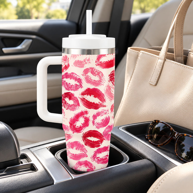 A tall handled tumbler with a white lid and straw sits in a car cup holder. The tumbler has a pink and red lip print pattern on a white background.