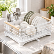 A white wire dish drying rack with light wood handles sits on a kitchen counter by a window, holding plates, bowls, cups, and utensils.