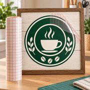 A framed coffee-themed graphic sign with a dark green circular logo featuring a white coffee cup and coffee beans, displayed on a wooden table next to a roll of pink-lined transfer or craft film.