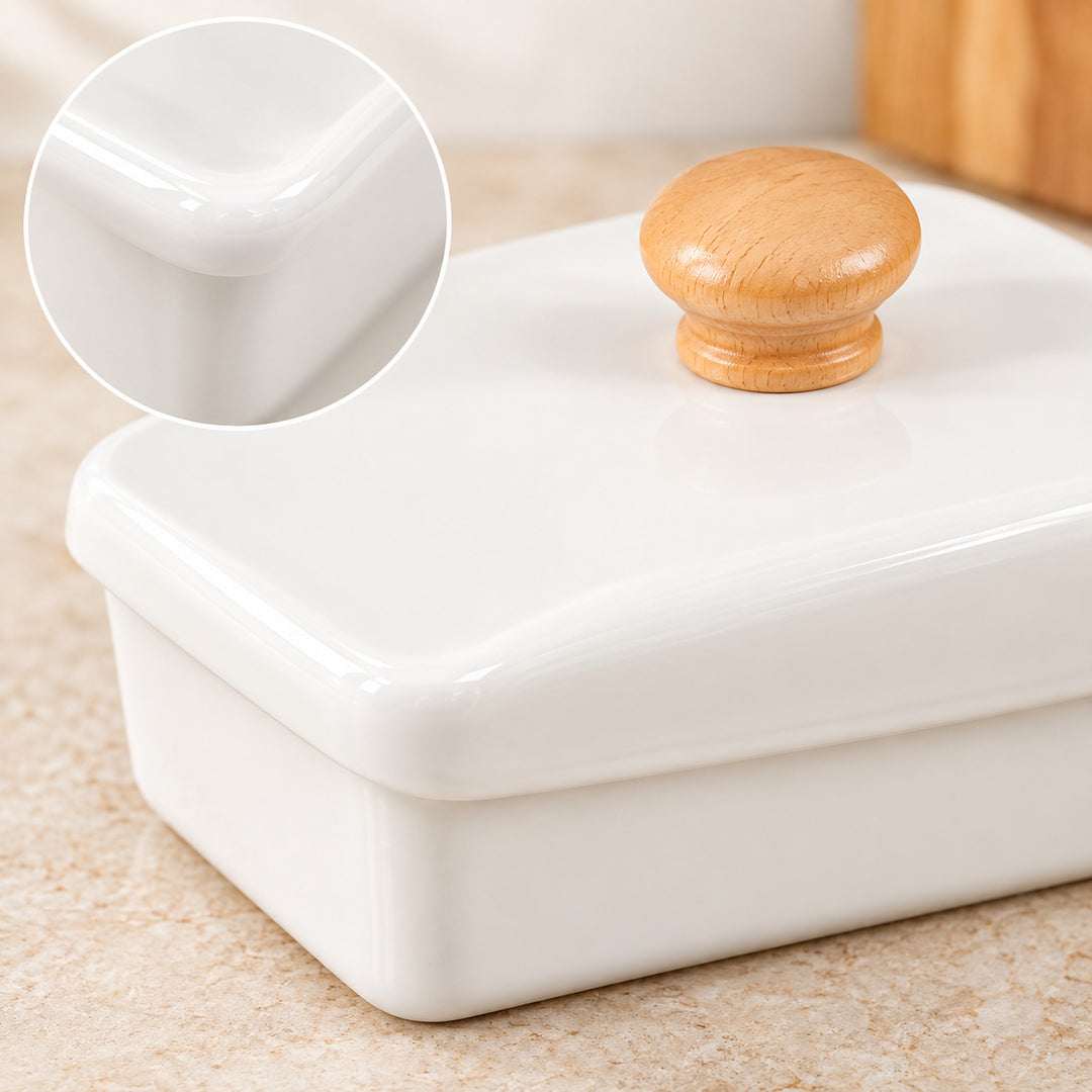 A glossy white ceramic butter dish with a rectangular shape and a round light wood knob on top, shown on a beige stone surface. A circular inset highlights the smooth, polished corner detail of the white ceramic.