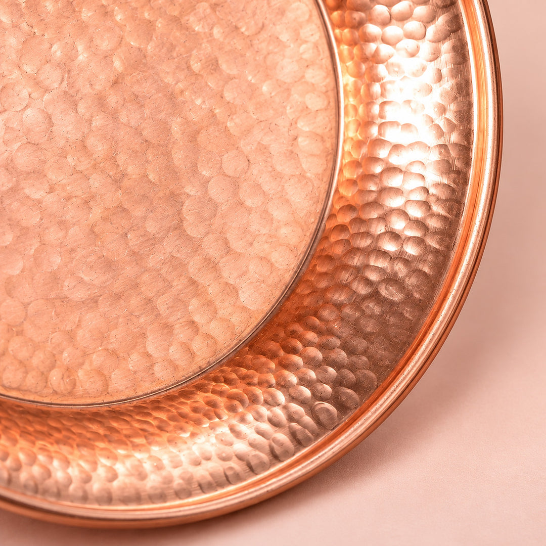 Close-up of a hammered copper tray or plate with a circular pattern, showing its metallic copper color and textured surface on a soft pink background.