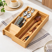 A bamboo utensil organizer tray with divided compartments holding metal forks, chopsticks, spoons, and wooden spoons, placed on a light countertop with a striped cloth and small kitchen props around it.