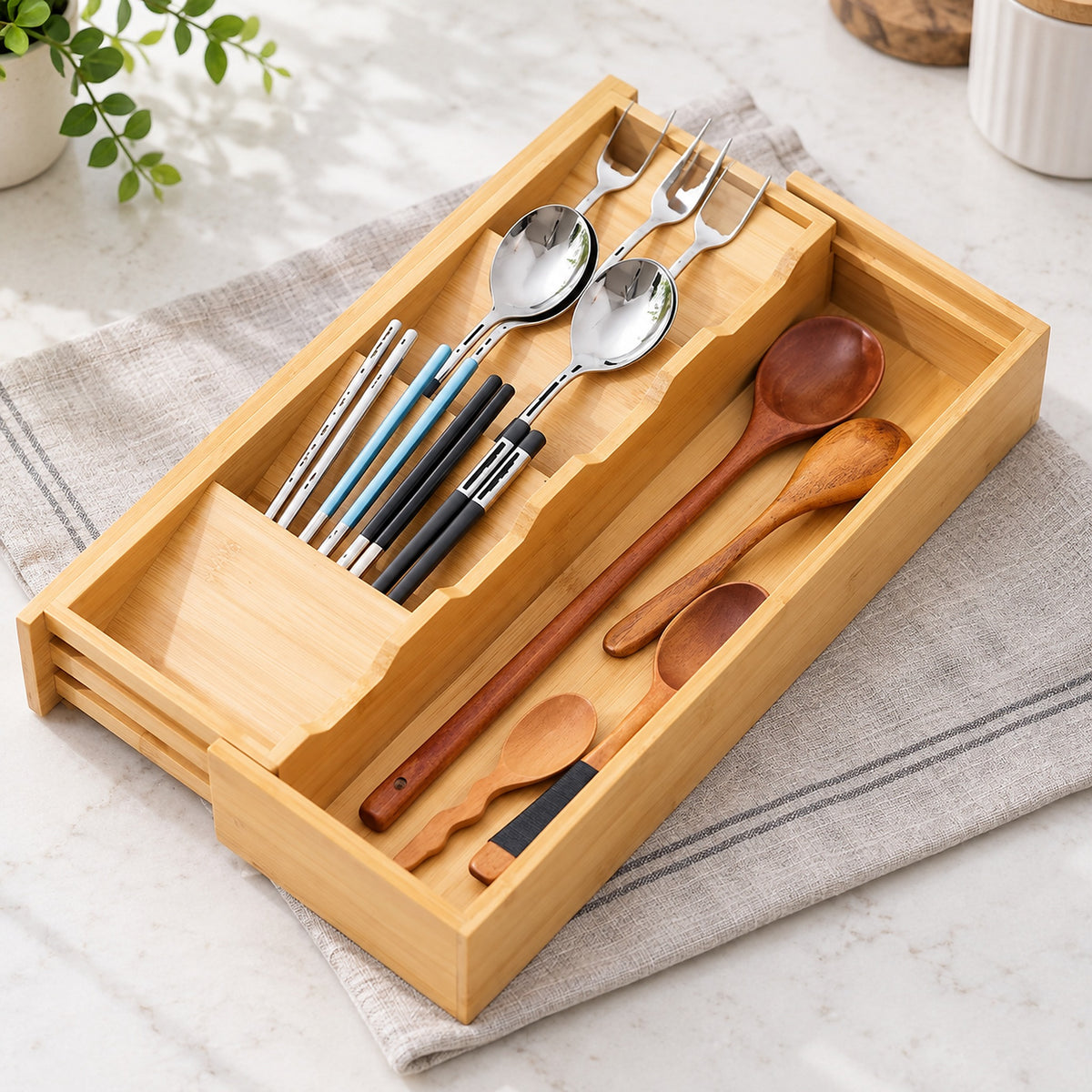 A bamboo utensil organizer tray holding assorted dining tools, including wooden spoons, wooden and metal chopsticks, and forks, placed on a folded beige cloth on a light stone surface.