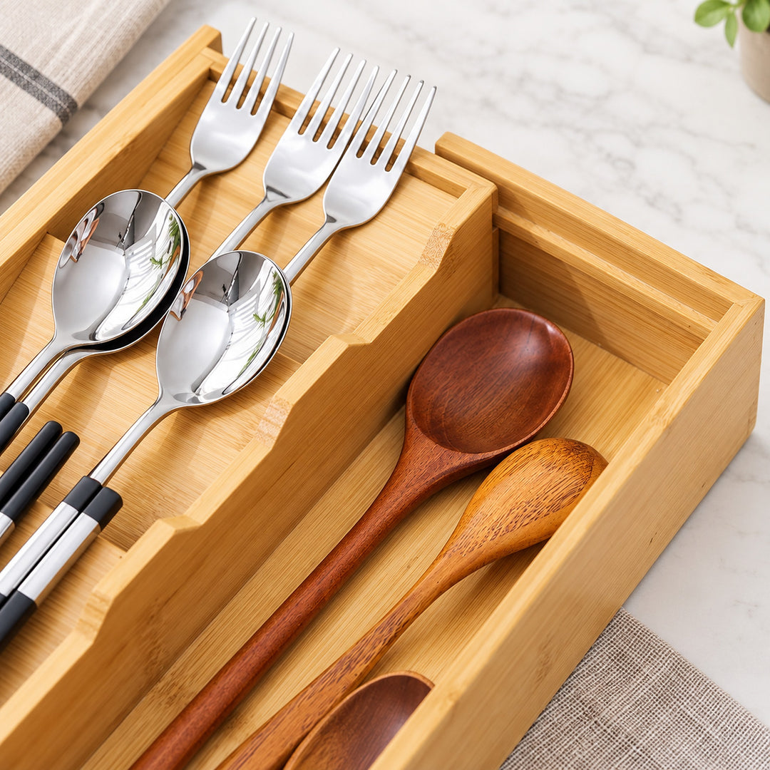A bamboo utensil organizer tray holding stainless steel spoons and forks on the left and two wooden spoons on the right, photographed on a light marble-like surface with a folded cloth in the corner.