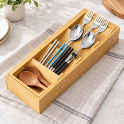 A bamboo utensil organizer tray holding silver spoons and forks, black chopsticks with blue accents, and two wooden spoons, placed on a striped beige cloth on a light marble surface.