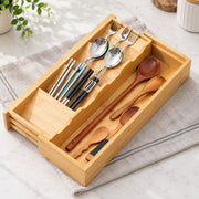 A bamboo utensil organizer tray holding assorted dining tools, including wooden spoons, wooden and metal chopsticks, and forks, placed on a folded beige cloth on a light stone surface.