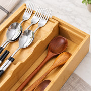 A bamboo utensil organizer tray holding stainless steel spoons and forks on the left and two wooden spoons on the right, photographed on a light marble-like surface with a folded cloth in the corner.