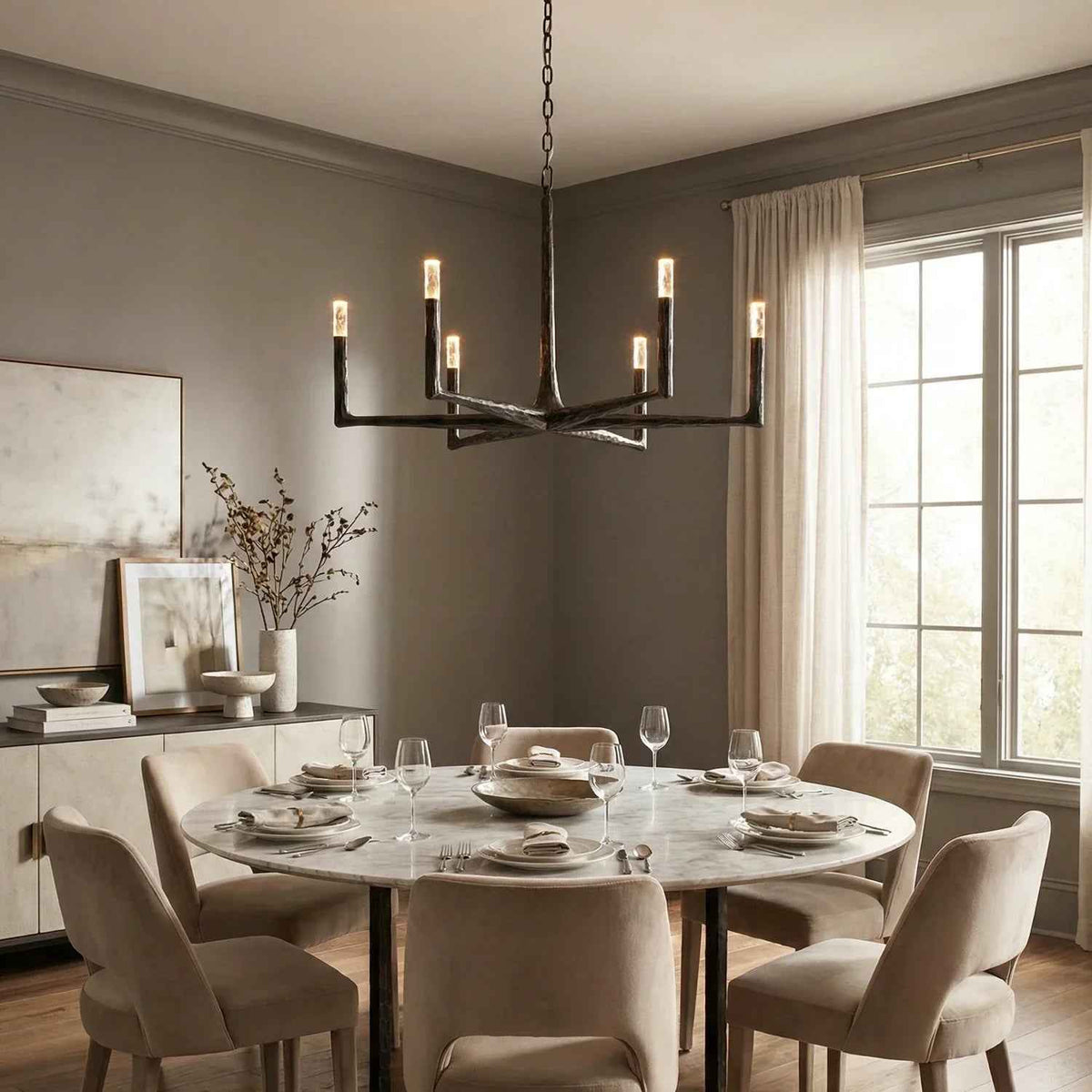 A modern multi-light chandelier with black metal arms and warm glowing Edison-style bulbs hangs over a round marble dining table set with beige upholstered chairs in a tastefully decorated dining room.