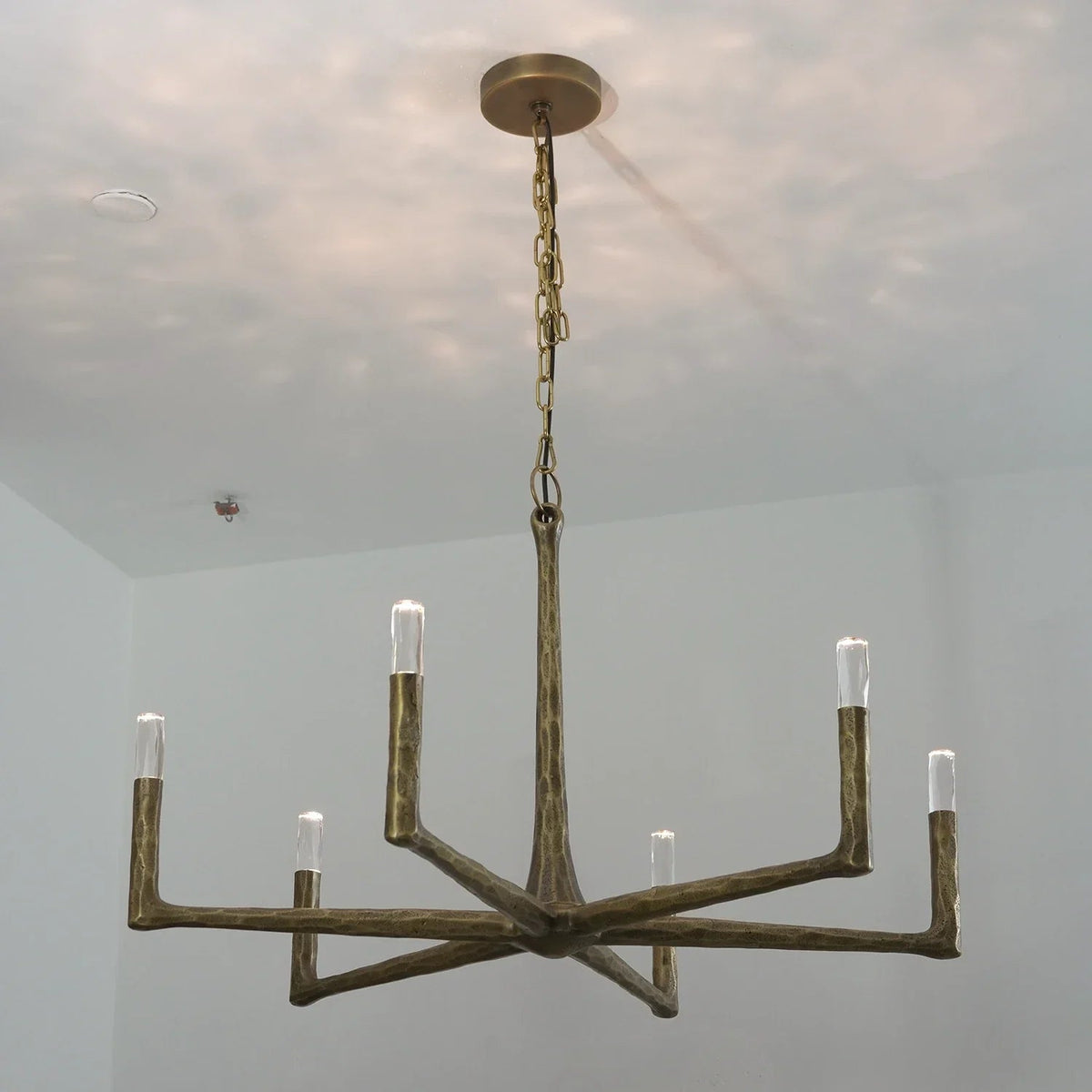 A rustic six-light chandelier with a distressed bronze finish, suspended from a ceiling by a chain, shown in a room with white walls.