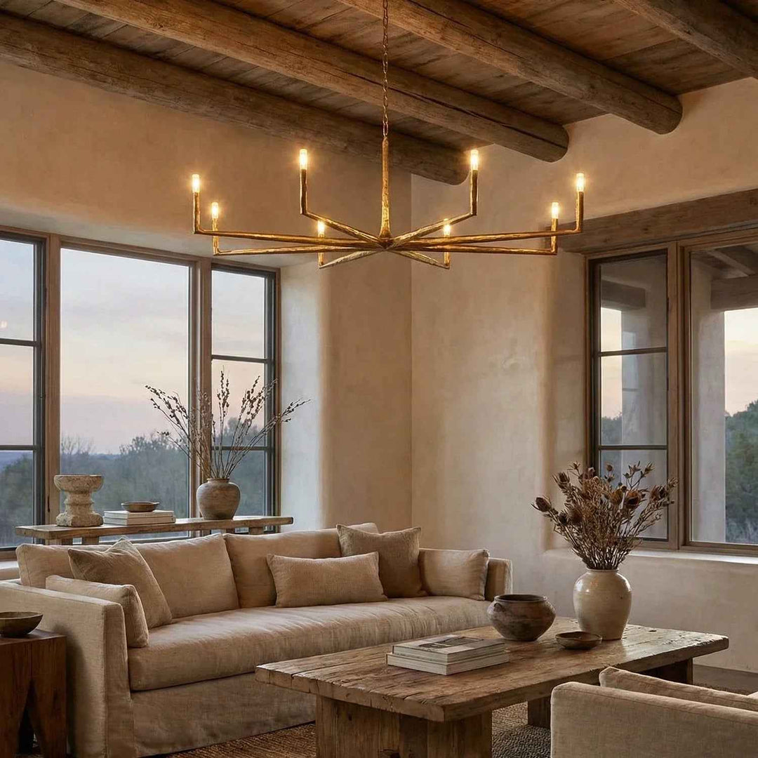 A modern brass multi-arm chandelier with candle-style bulbs hangs in a rustic, warm-toned living room with a beige sofa, wooden ceiling beams, and large windows.