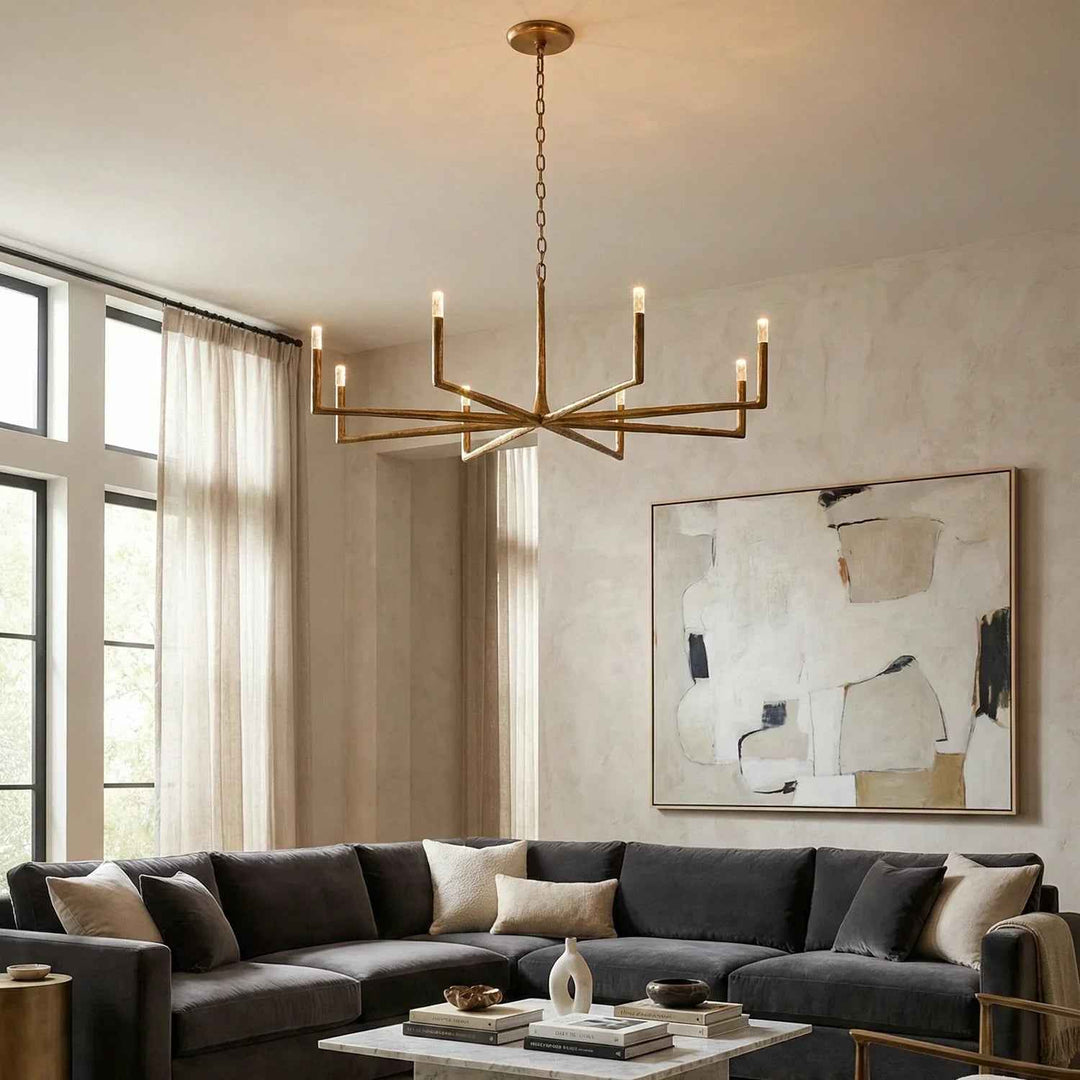 A modern multi-light chandelier with a gold-finished frame and candle-style bulbs hangs in a styled living room above a dark gray sectional sofa. The room features textured beige walls, a large abstract painting, and tall windows with sheer curtains.