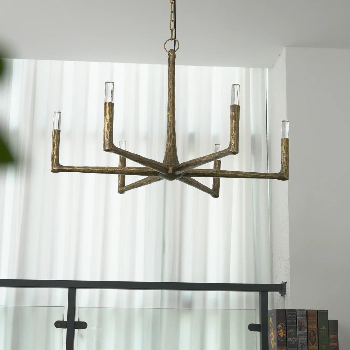 A rustic five-light branch-style chandelier with a distressed bronze finish hangs in a modern room with white curtains and a glass railing.