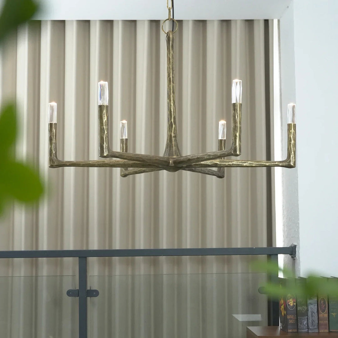 A modern six-light chandelier with a textured, branch-like metallic frame in a warm gold finish, hanging in front of beige pleated curtains.