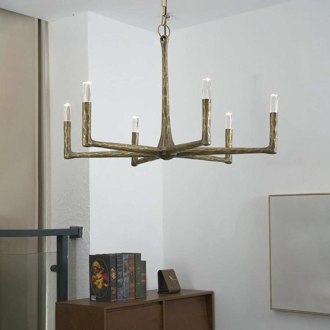 A modern six-light chandelier with a textured, branch-like metallic gold finish hangs in a minimalist room with white walls and a wooden console table below.