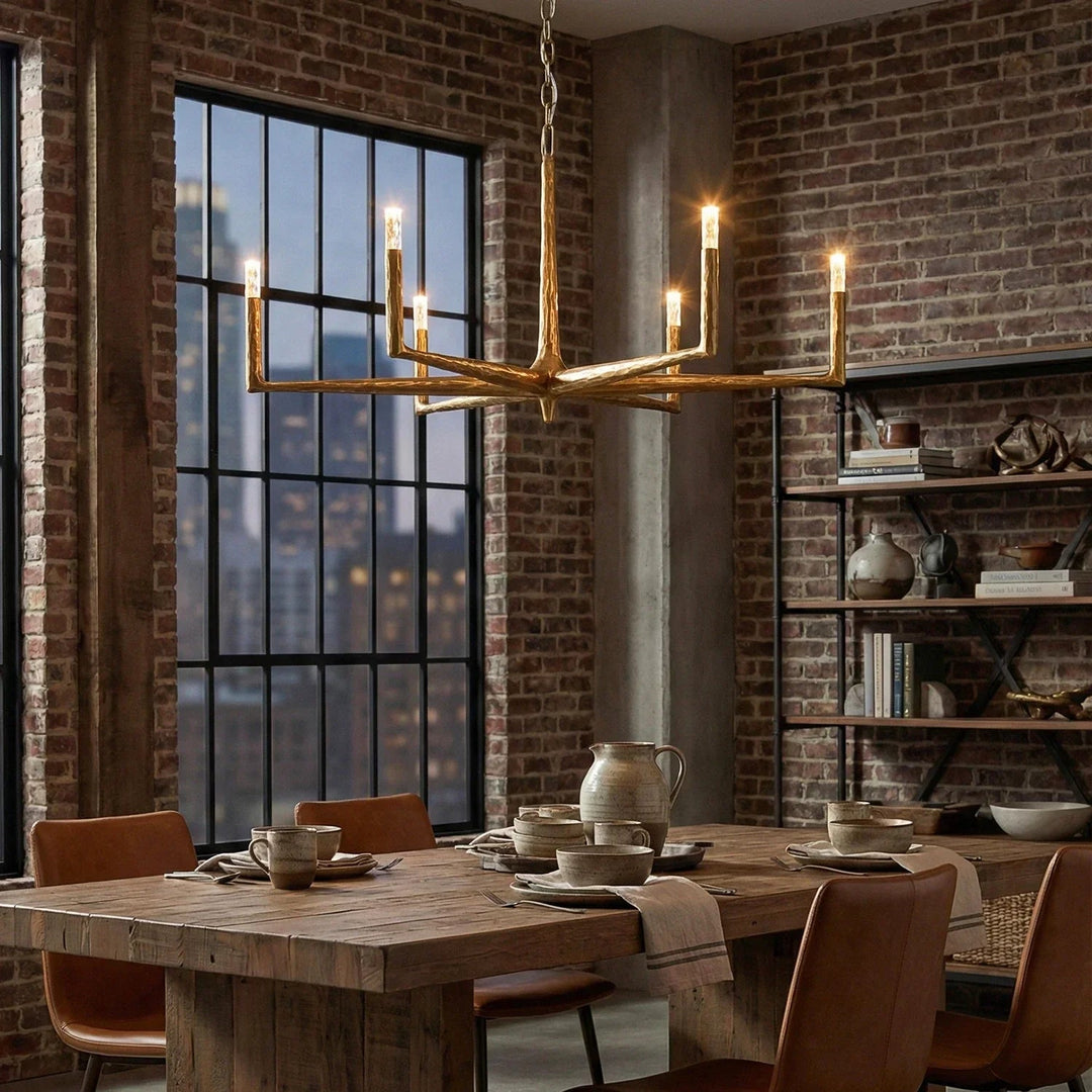 A modern brass multi-light chandelier with candle-style bulbs hangs over a rustic wooden dining table in a loft-style room with exposed brick walls, black metal shelving, and large industrial windows.