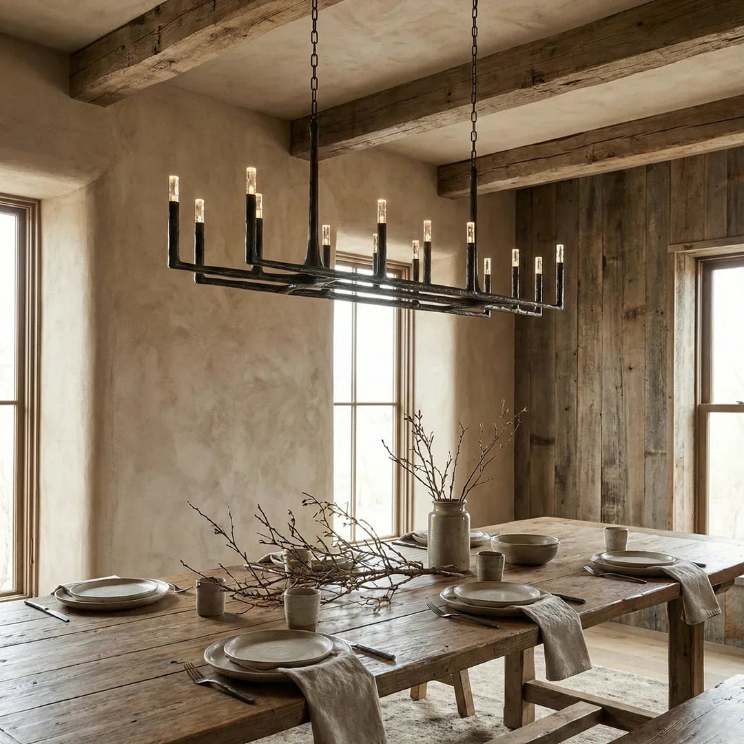 A rustic black metal multi-light chandelier with candle-style bulbs hangs over a long wooden dining table set with plates and neutral decor in a warm, wood-paneled room.
